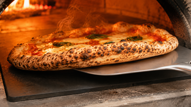 Carbon steel pizza stone lifting a leopard-spotted pizza with a crisp blistered base.
